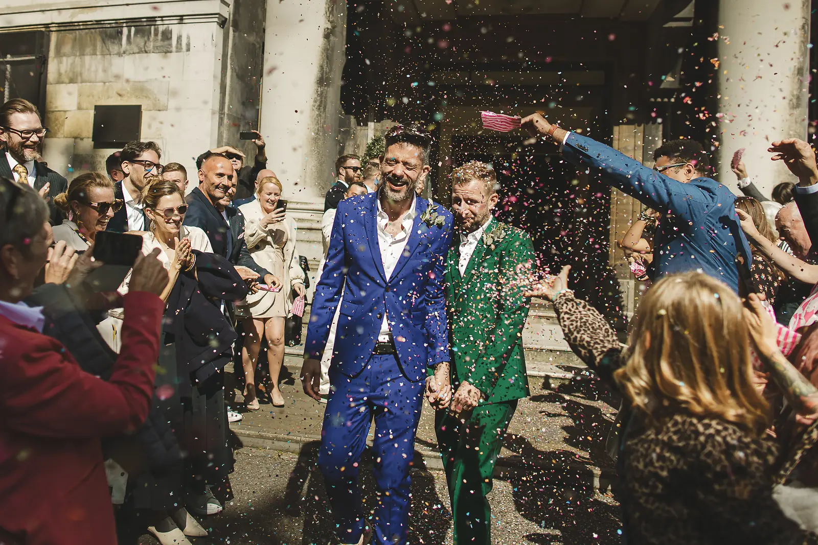 Two grooms walking through a colourful confetti shower outside a London registry office, one in a cobalt blue suit and one in an emerald green suit, guests cheering around them
