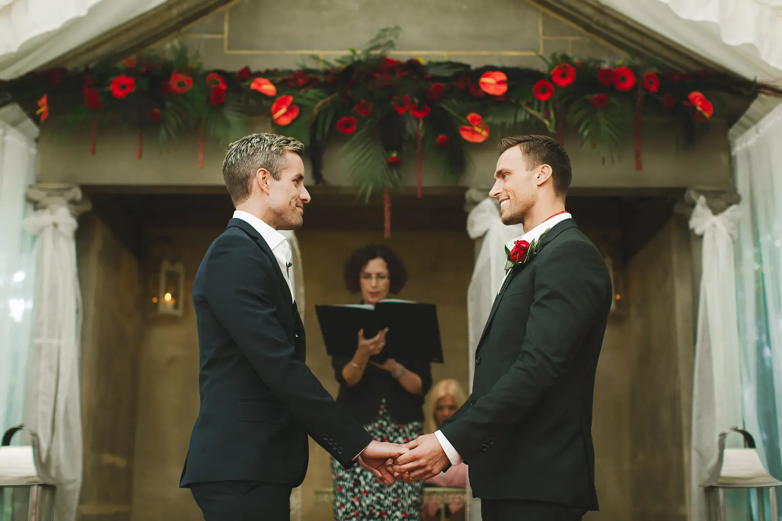 Two grooms holding hands and exchanging vows at Parley Manor Dorset beneath a dramatic garland of red anthurium flowers and tropical foliage, officiant reading behind them