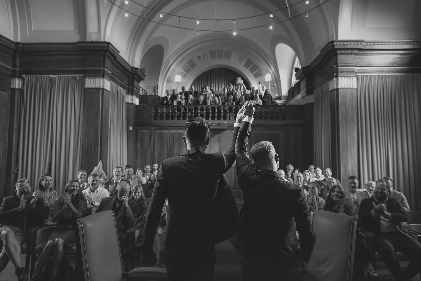 Black and white image of two grooms seen from behind raising their clasped hands in triumph as guests in a grand domed London ceremonial hall rise to applaud