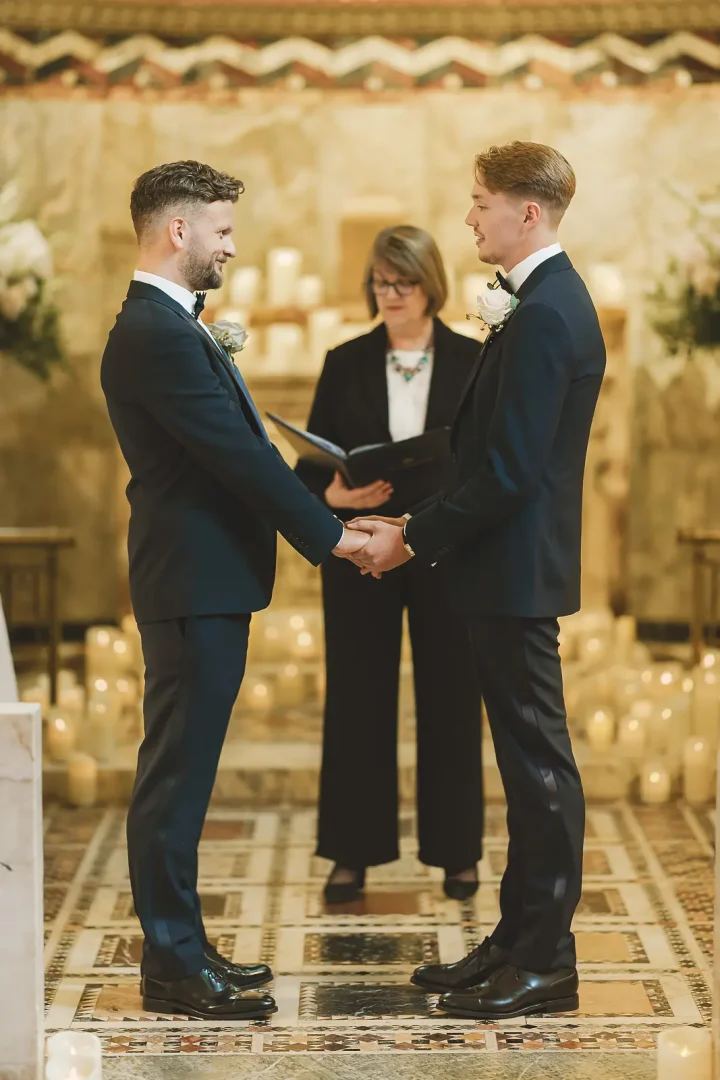 Two grooms holding hands and smiling during their wedding ceremony in a grand candlelit marble hall with hundreds of glowing pillar candles and officiant behind them