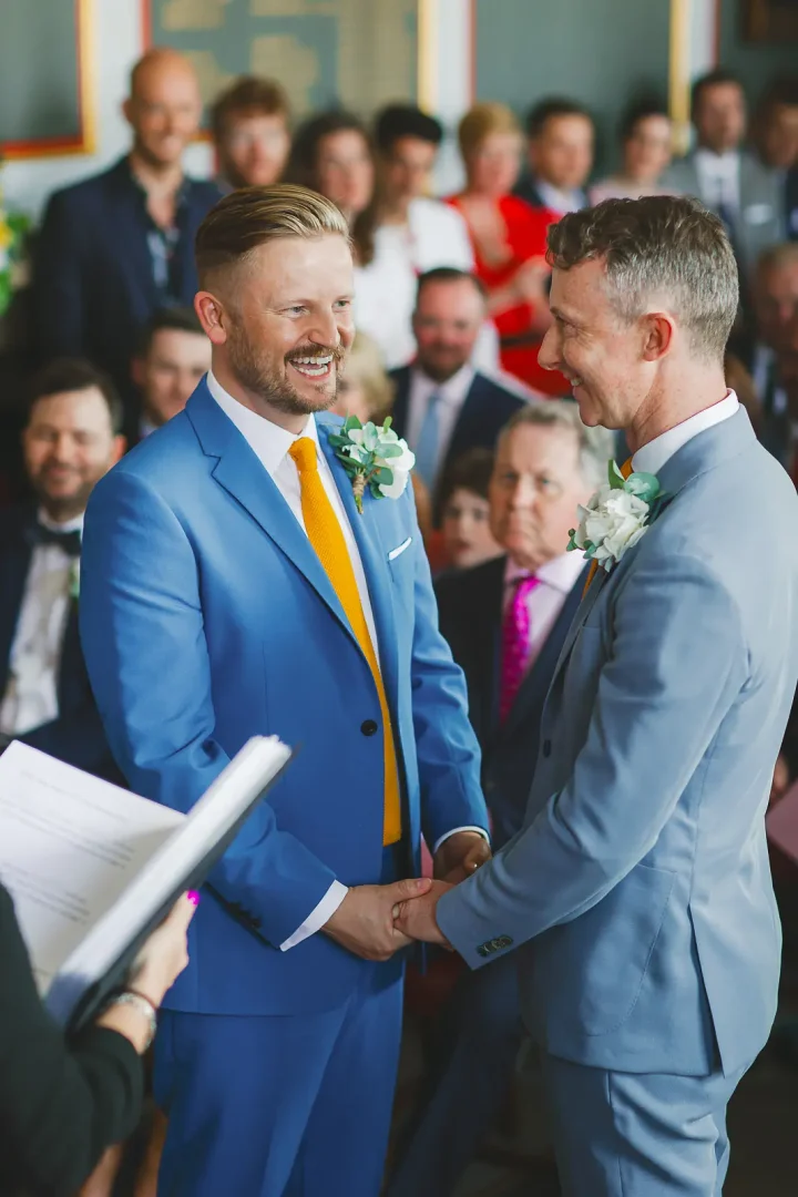 Two grooms in colourful suits smiling and holding hands during their wedding ceremony as guests look on with joy