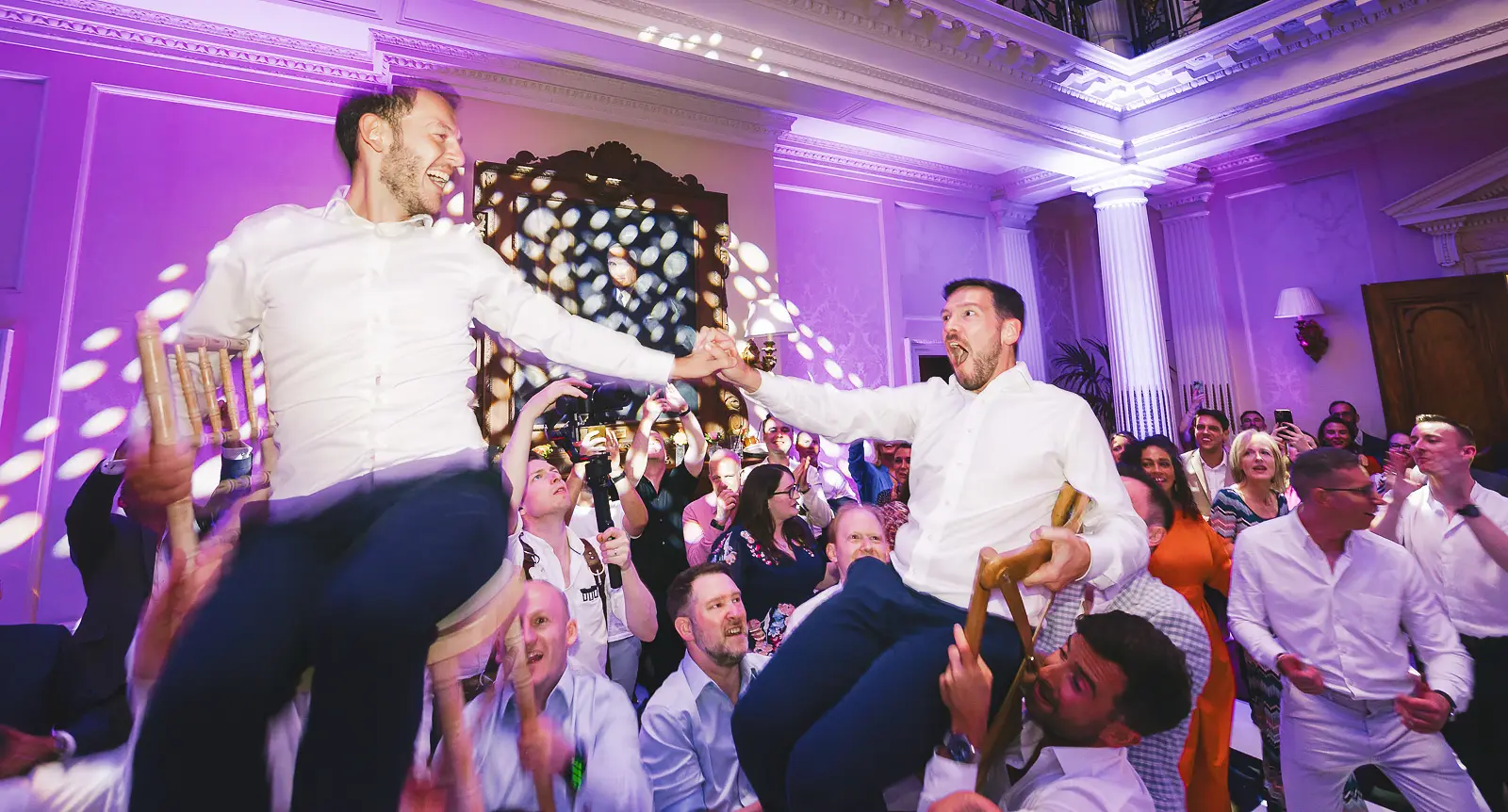 Two grooms reaching toward each other from raised chairs during the Hora dance at their Jewish wedding reception, surrounded by joyful cheering guests in a grand ballroom lit in purple