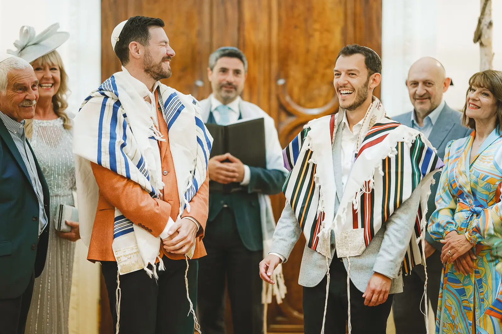 Two grooms laughing with joy during their Jewish wedding ceremony, both wearing colourful tallits and kippot, with family and the officiant gathered around them