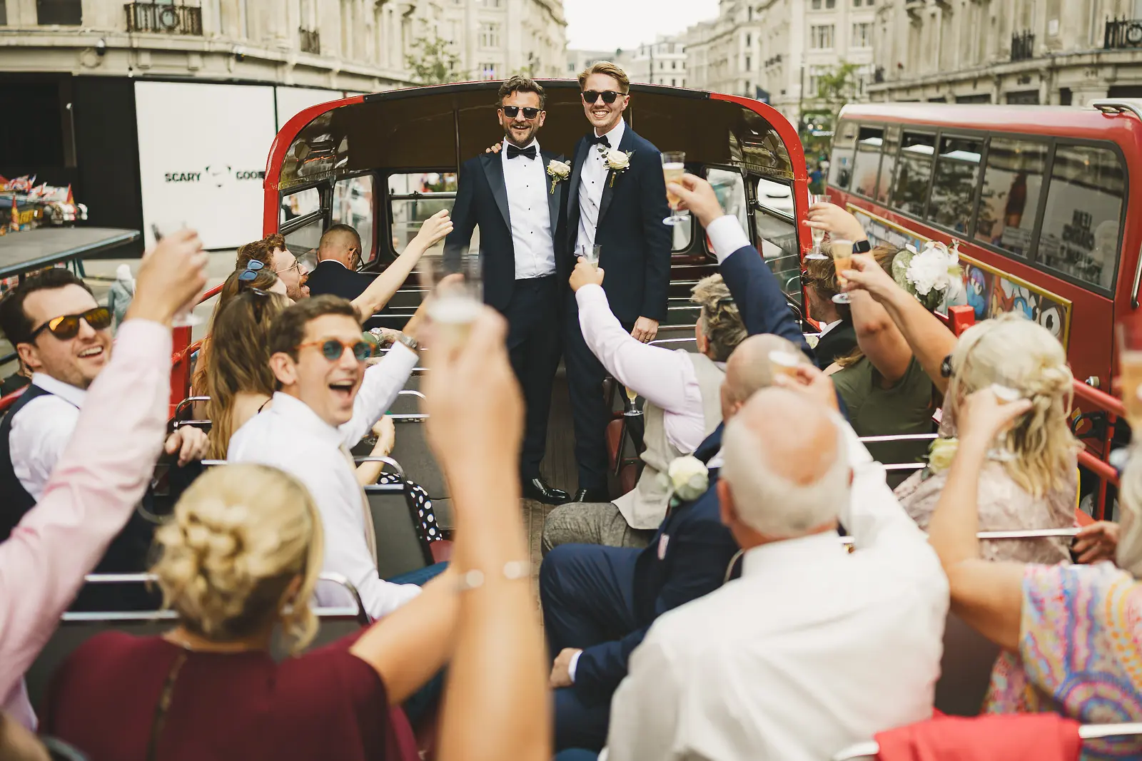 Two grooms in navy tuxedos and sunglasses standing on an open-top red London bus as wedding guests raise champagne glasses and cheer along Regent Street behind them