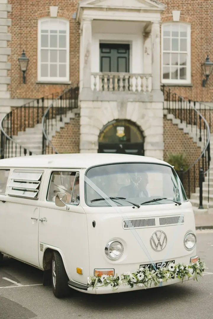 Vintage white VW campervan decorated with daisy garland parked outside Poole Guildhall Registry Office with grand staircase behind