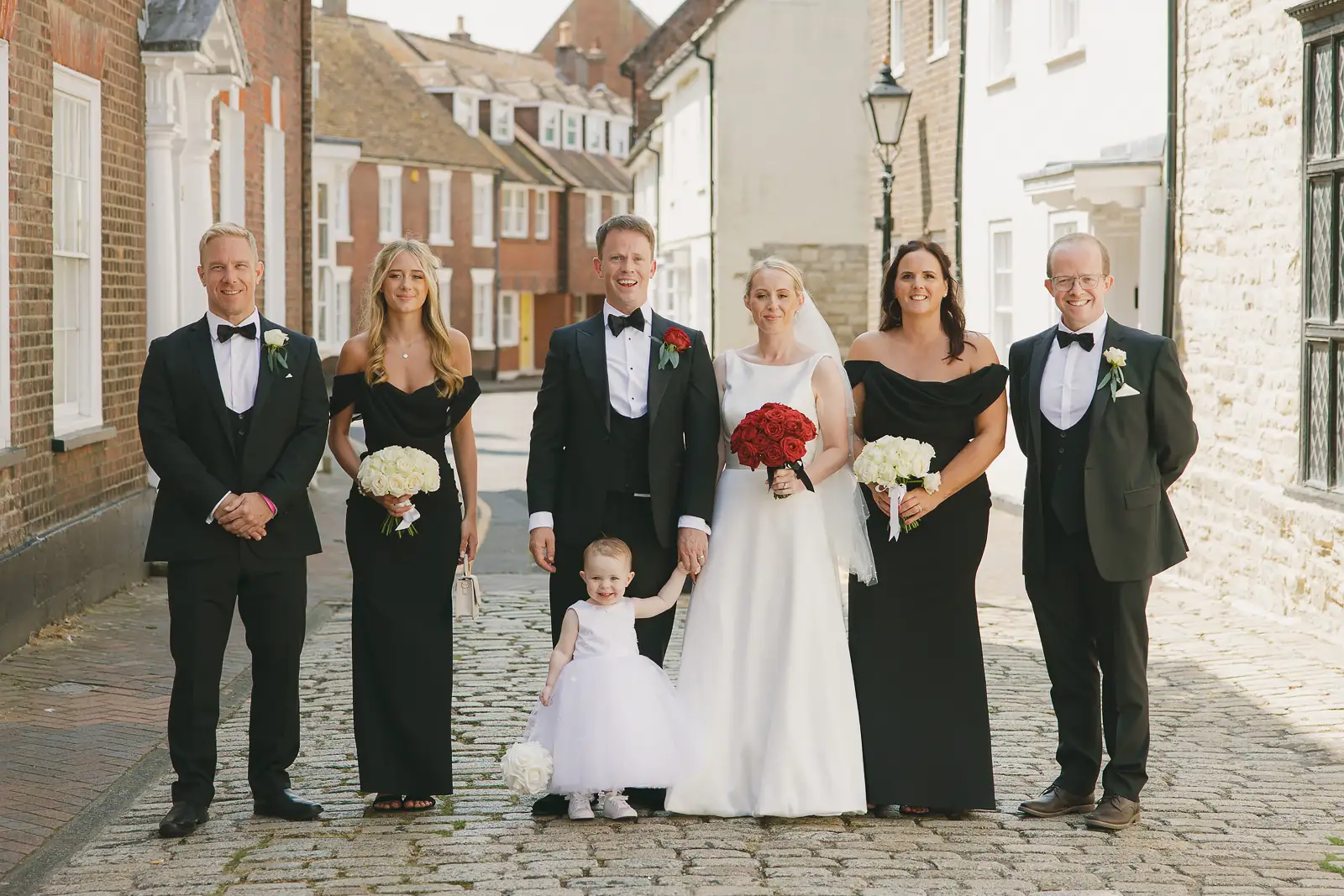 Wedding party in black tie with bridesmaids and flower girl posing on a cobbled street in Poole Old Town after Poole Guildhall registry office wedding, red and white bouquets