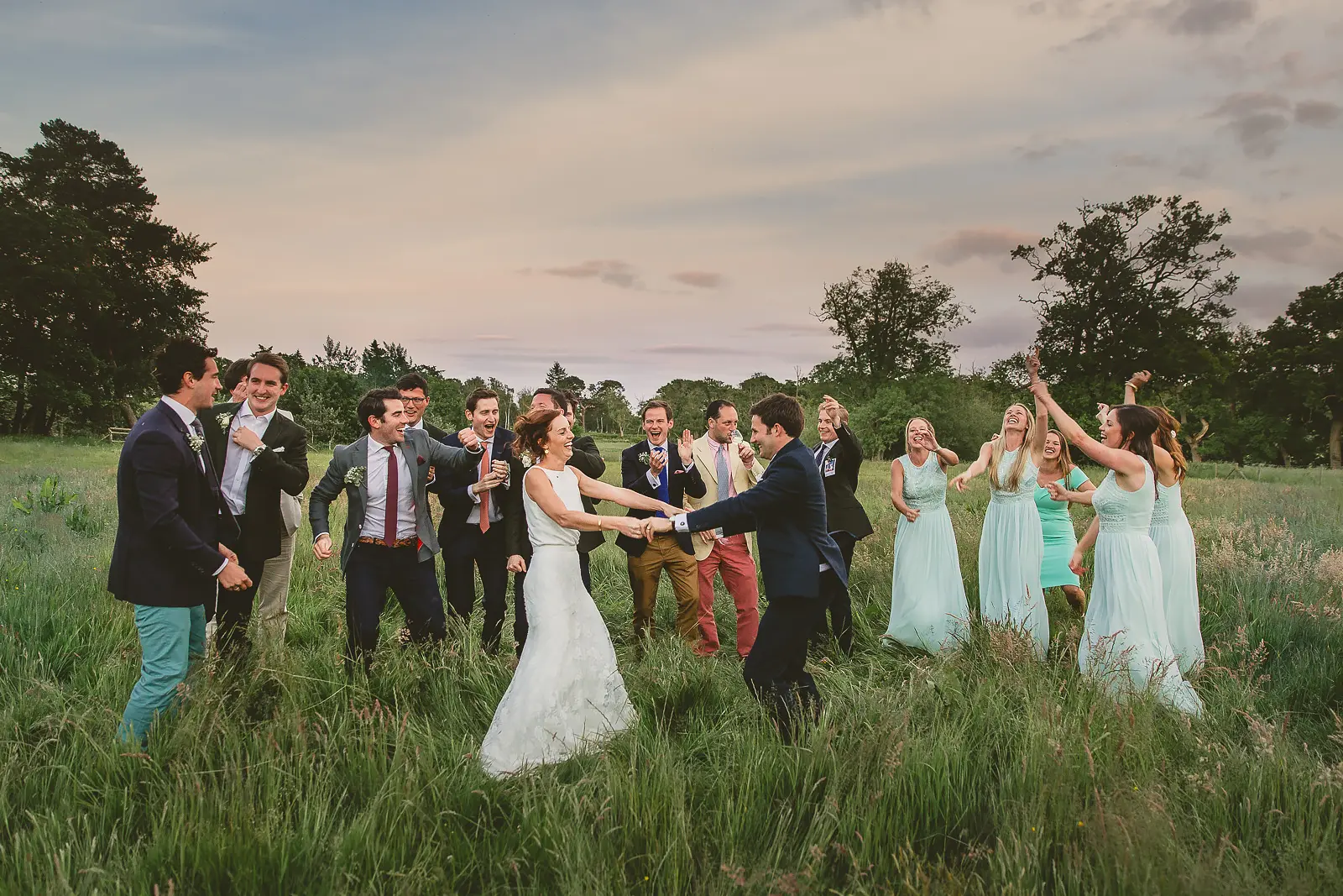 Bride, groom and wedding party dancing and celebrating freely in an open Dorset field at dusk, colourful outfits against a wide dramatic evening sky