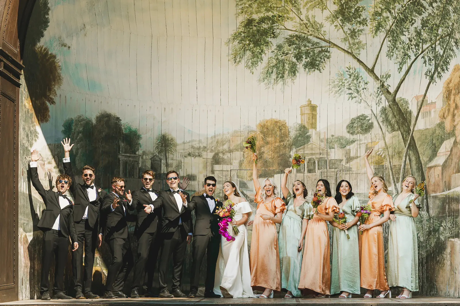 Wedding party celebrating on the outdoor theatre stage at Larmer Tree Gardens, groomsmen in black tie and bridesmaids in peach and sage dresses with arms raised before a grand painted scenic backdrop