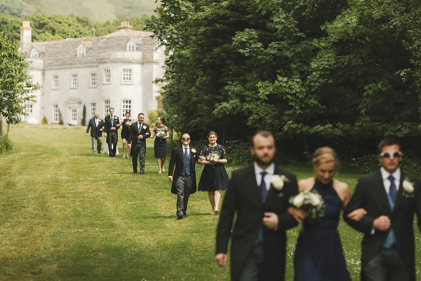 Wedding party walking in procession across the lawn at Smedmore House Dorset with the white Georgian manor and wooded hillside visible in the background