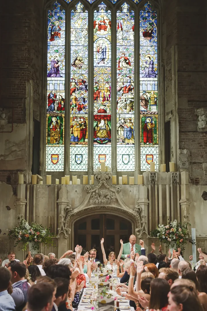 Wedding guests raising hands cheering during a toast in a grand gothic banquet hall dominated by a vast ornate stained glass window and candlelit altar