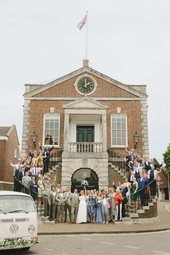 Entire wedding party celebrating on the steps of Poole Guildhall with Union Jack flying and vintage VW campervan in foreground