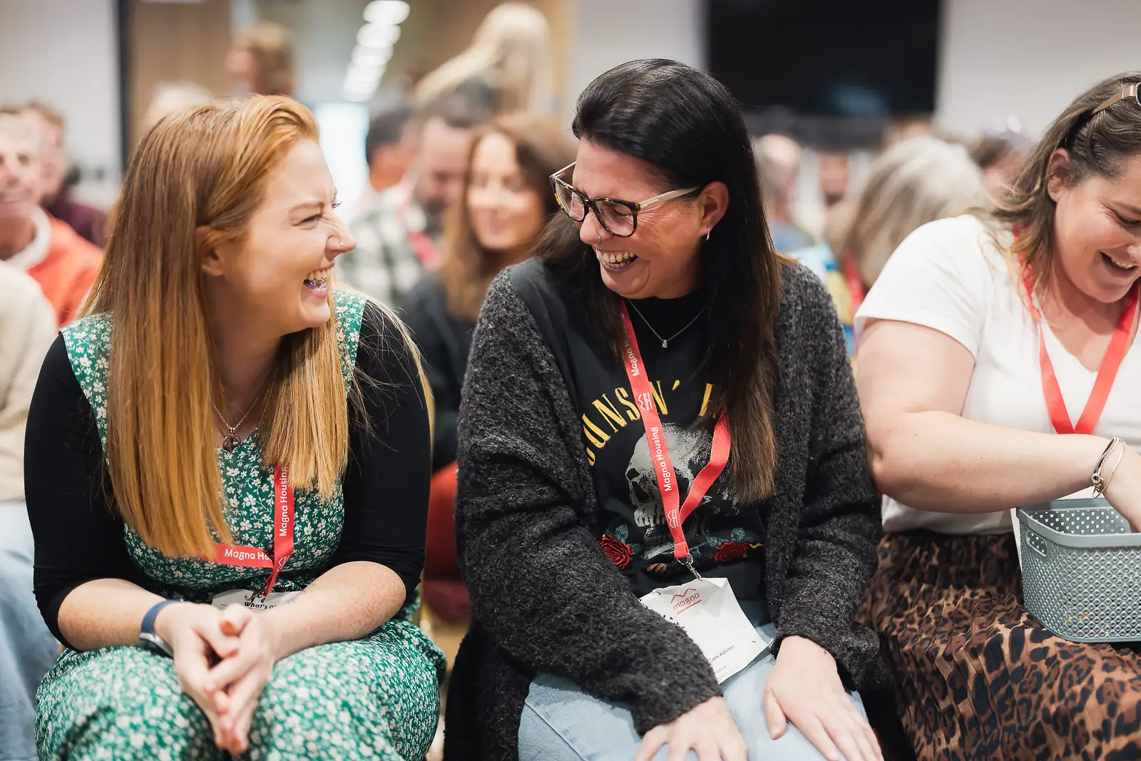 Two colleagues laughing together in the audience during a session at a corporate away day conference, wearing branded lanyards