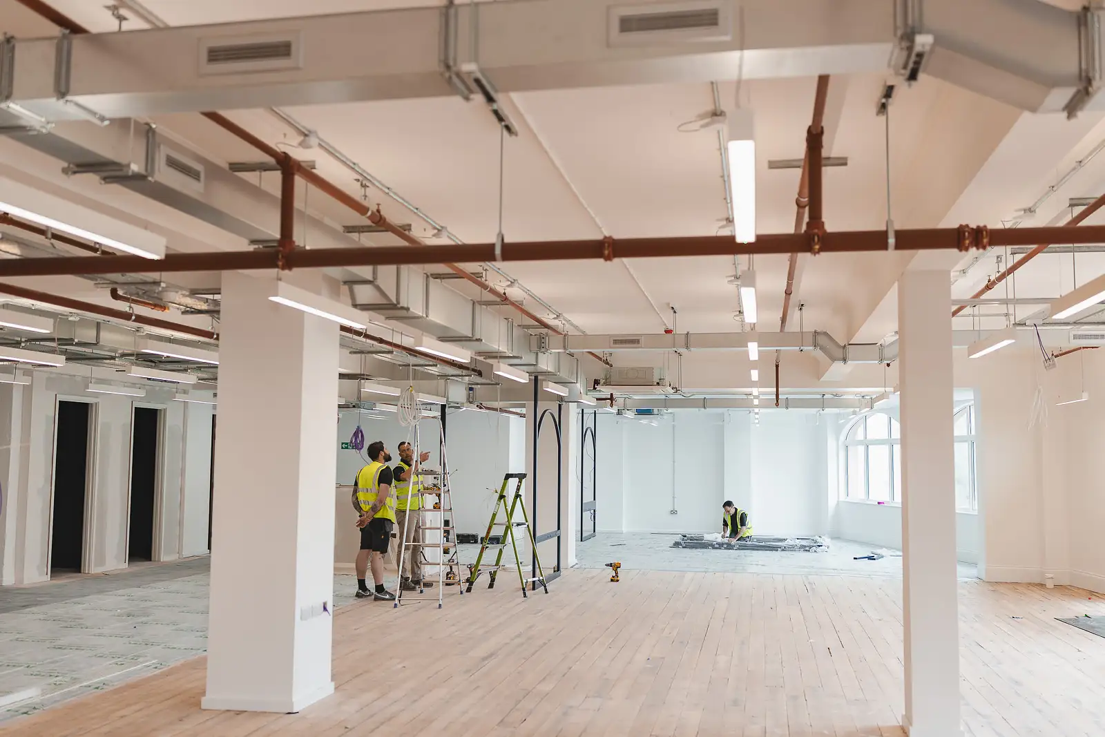 Wide floor-plate shot of construction team at work, copper pipe services running overhead — commercial photographer on location