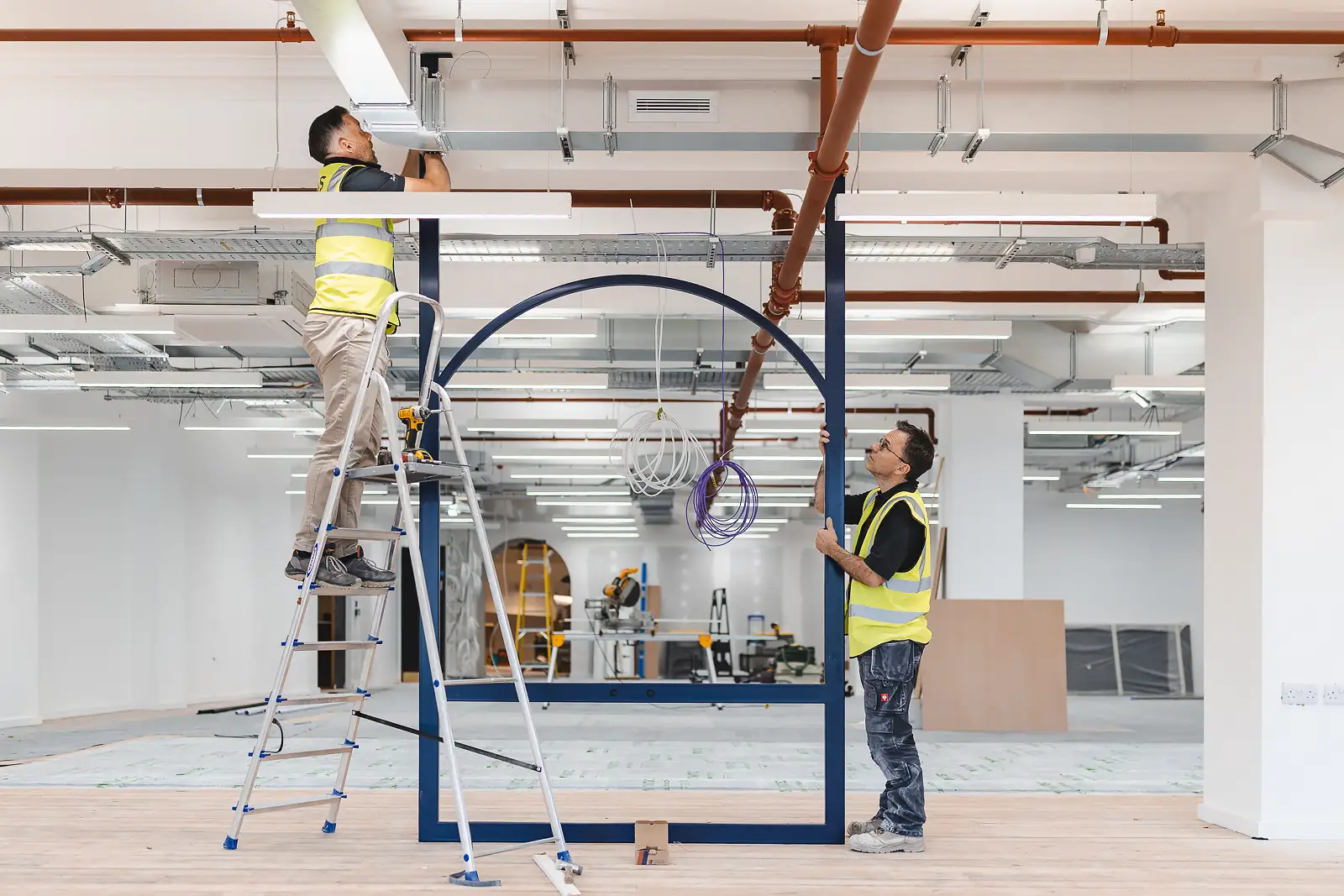 Two workers lifting and positioning a steel arch frame in an open-plan floor, wide perspective by commercial photographer