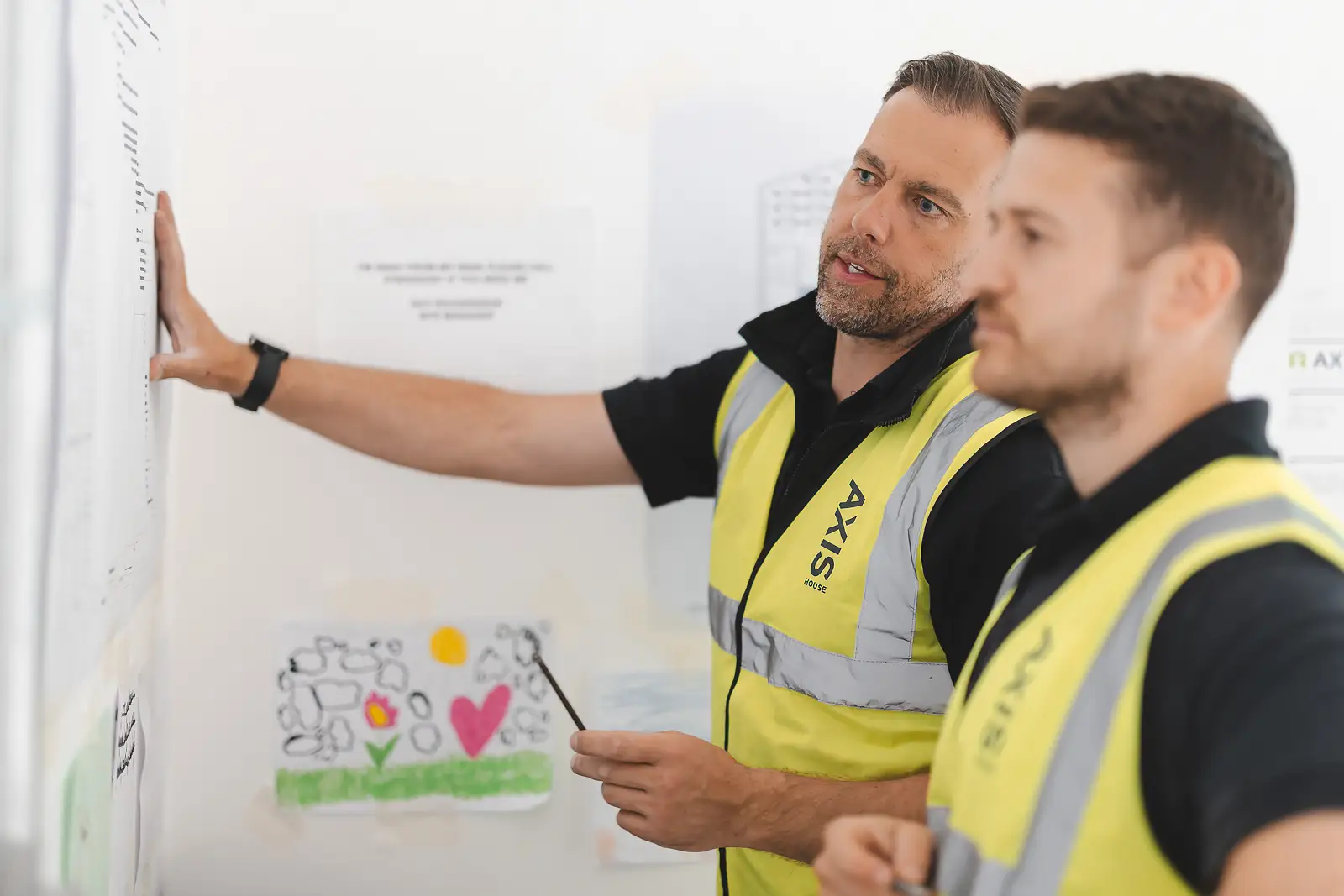 Two site managers reviewing plans pinned to the wall, senior manager pointing out key details — commercial photography on location