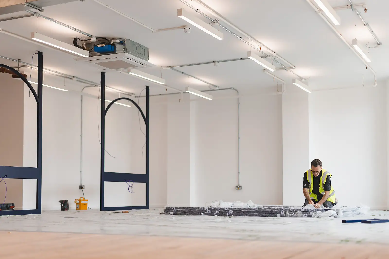 Wide shot of open-plan office fitout in progress, steel arch frames in place, worker kneeling with flooring tools — commercial photography