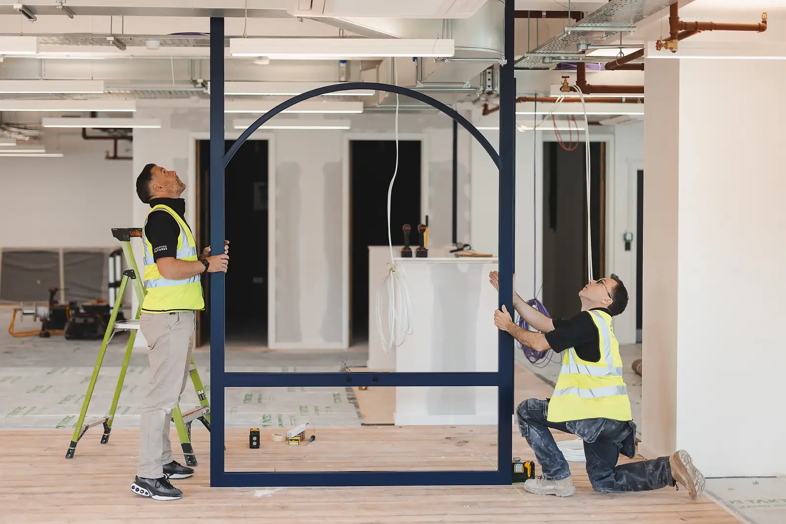 Two workers installing a large steel arch frame, wide-angle shot showing scale of the space — commercial photography on site