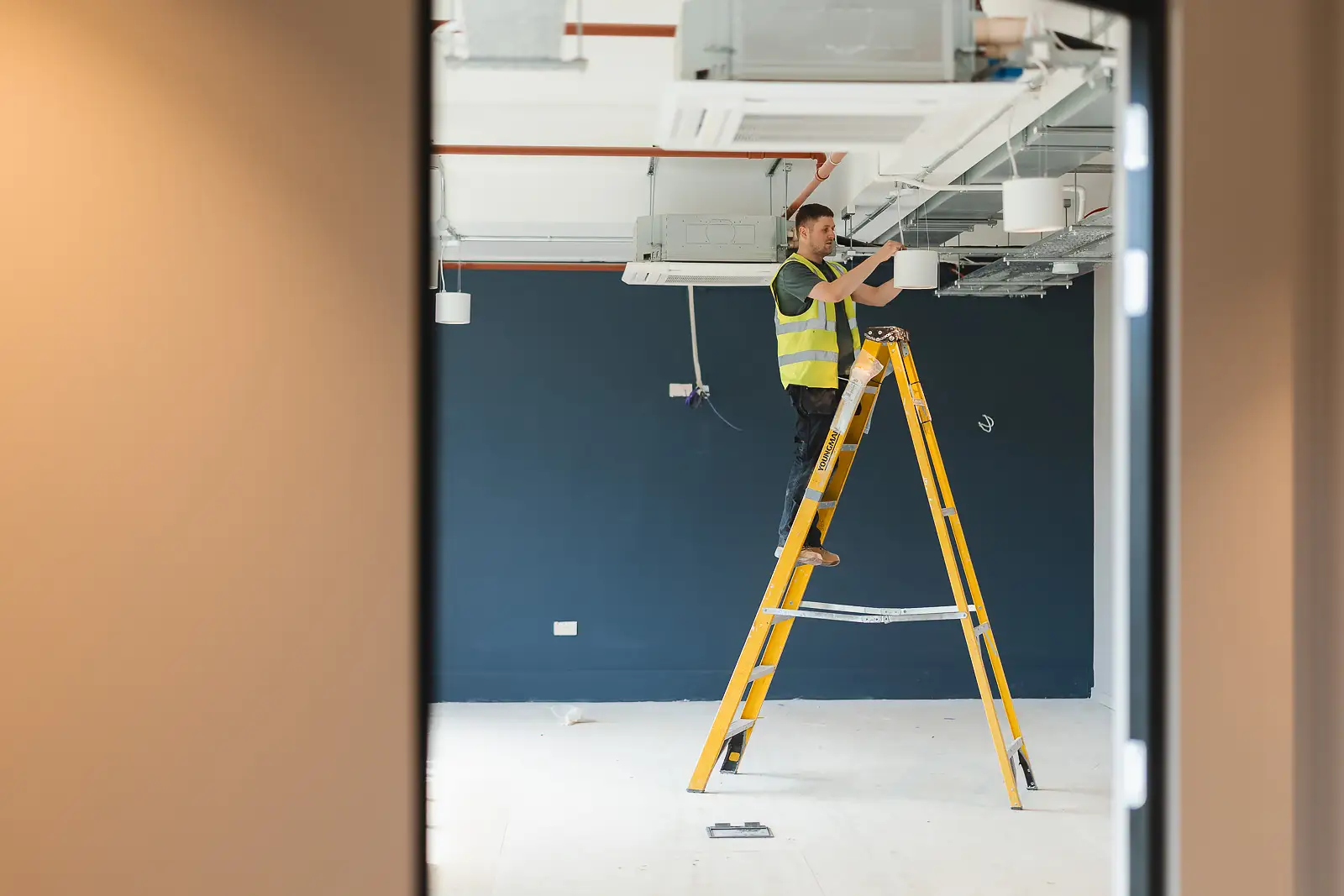 Electrician on a yellow ladder working at ceiling height, photographed through a doorway frame into a navy-painted office