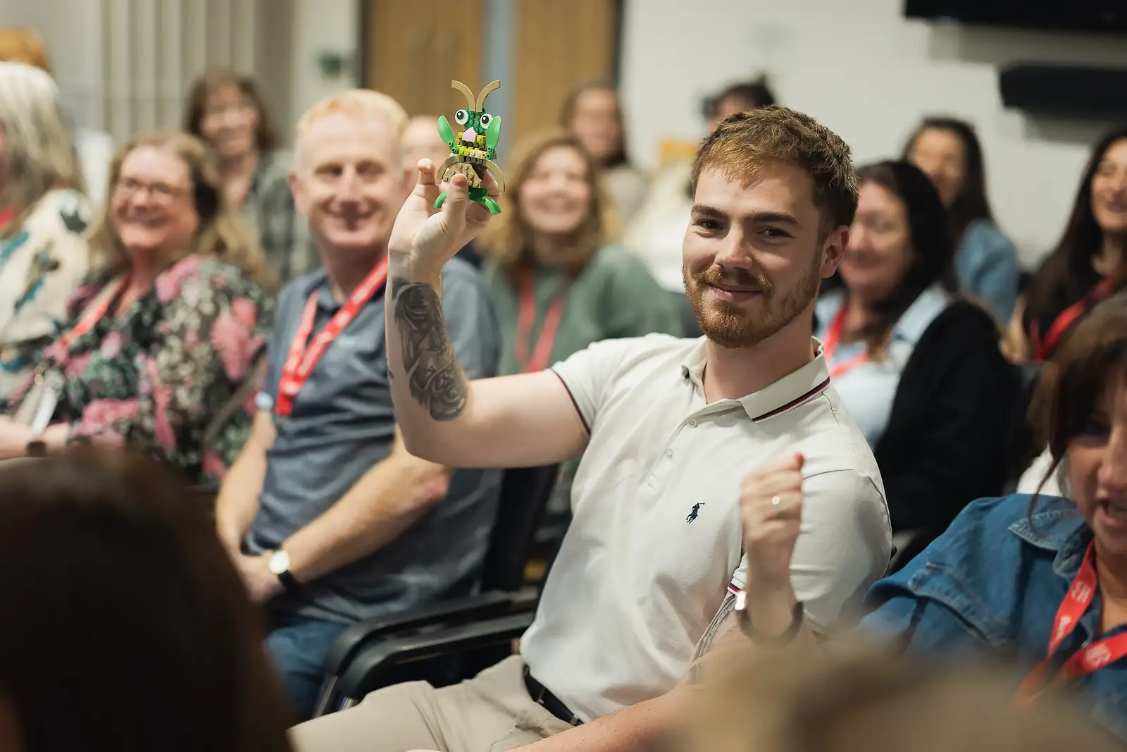 Smiling employee proudly holds up a small Lego model to laughing colleagues during a fun moment at a corporate event