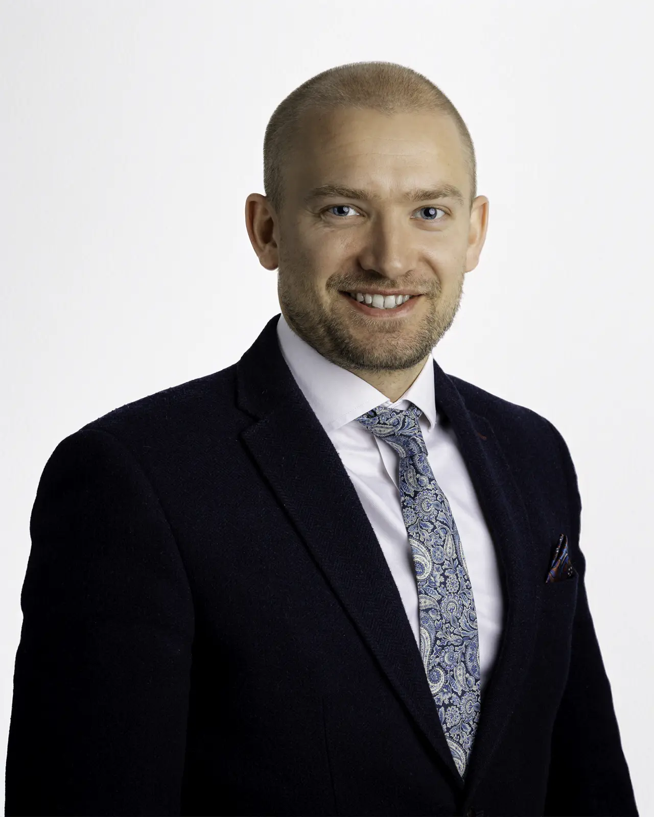 Professional executive headshot of a man in a navy suit and paisley tie against a light background