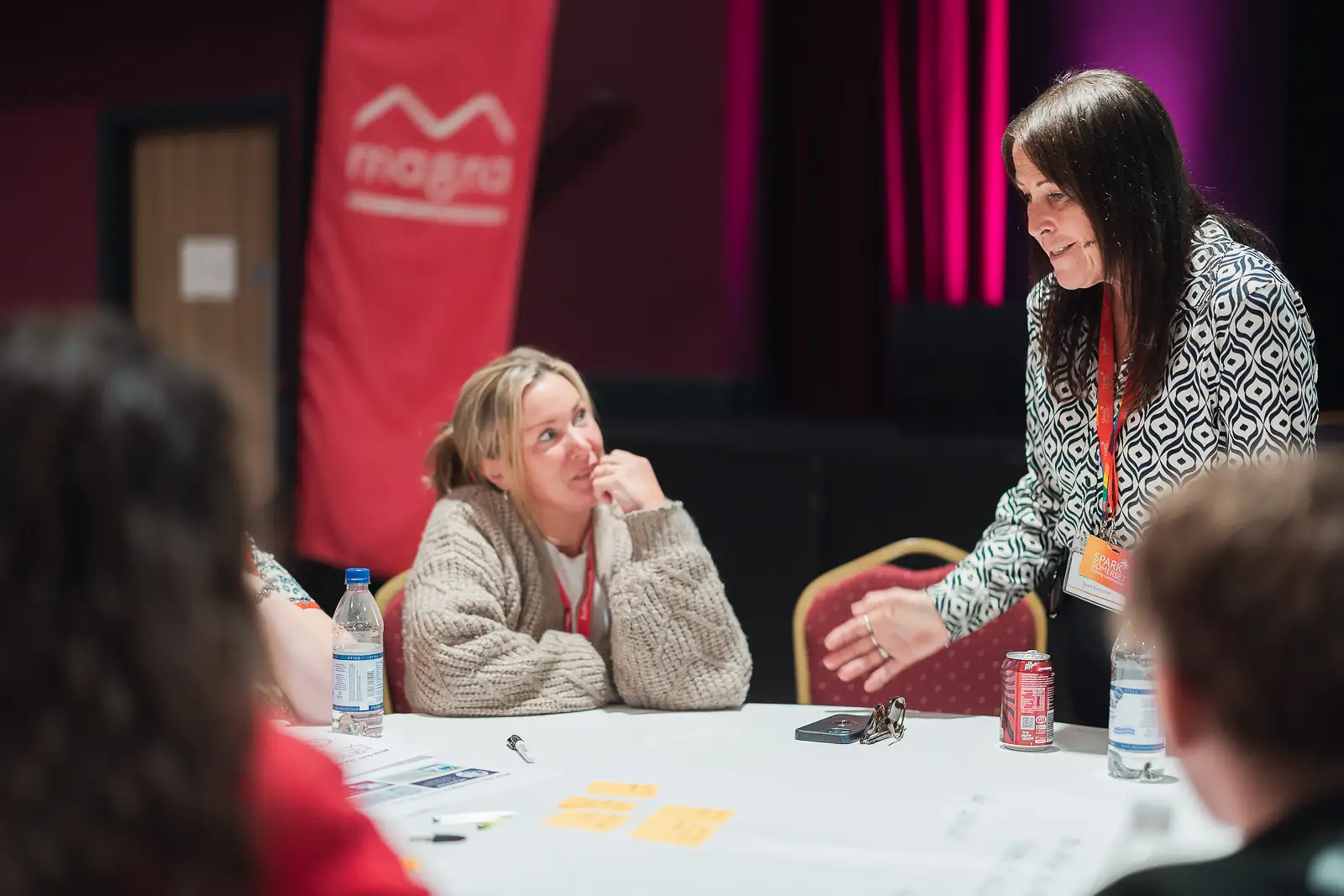 Facilitator leading a table discussion with engaged colleagues at a corporate conference in Somerset, branded banner visible in background