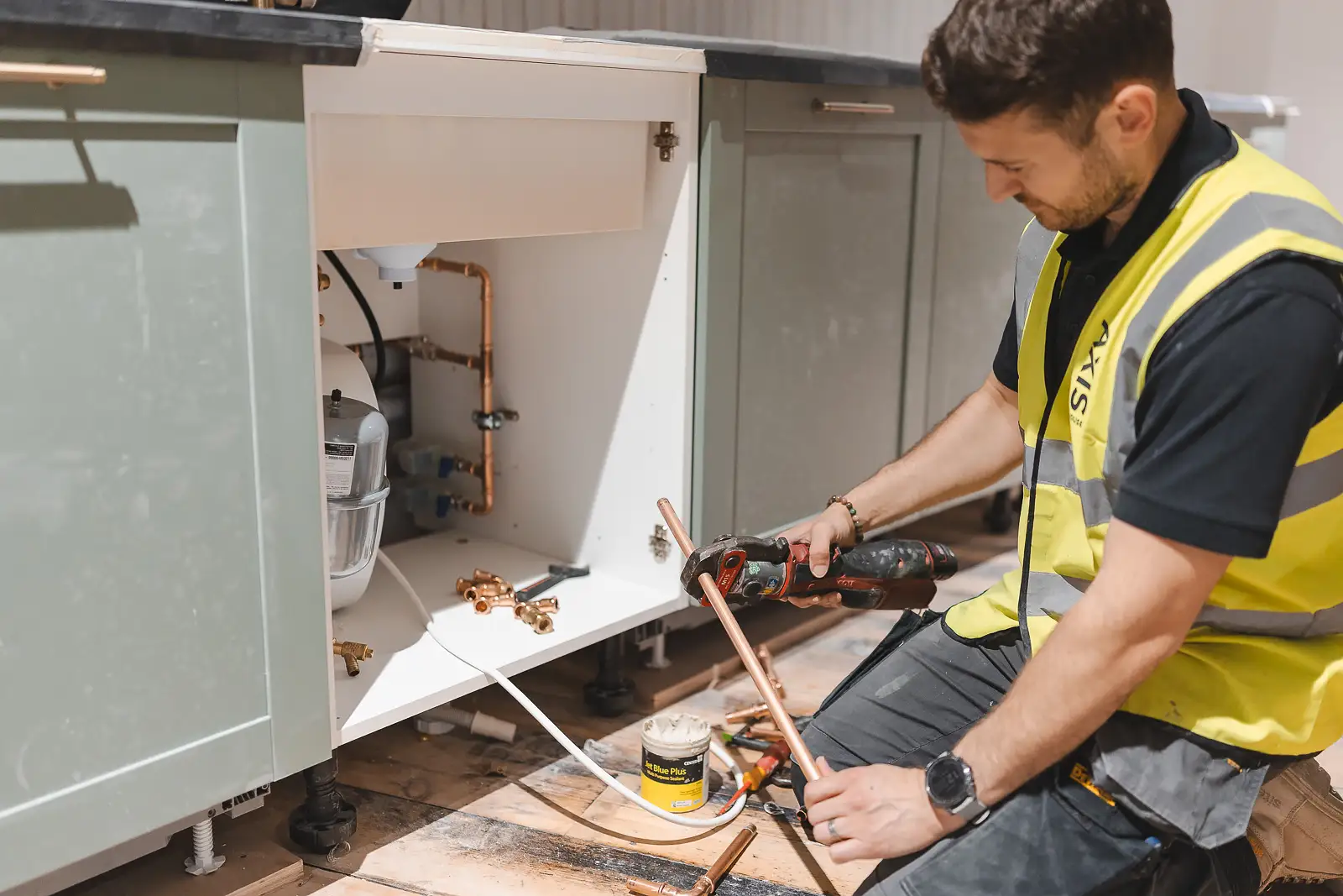 Plumber using a pipe press tool to fit copper pipework beneath a sage green kitchen unit — lifestyle photography in Bournemouth