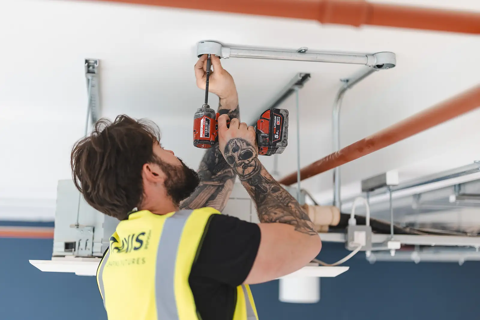 Low-angle shot looking up at a tradesman with tattooed arms using a Milwaukee cordless drill overhead on conduit — lifestyle photography
