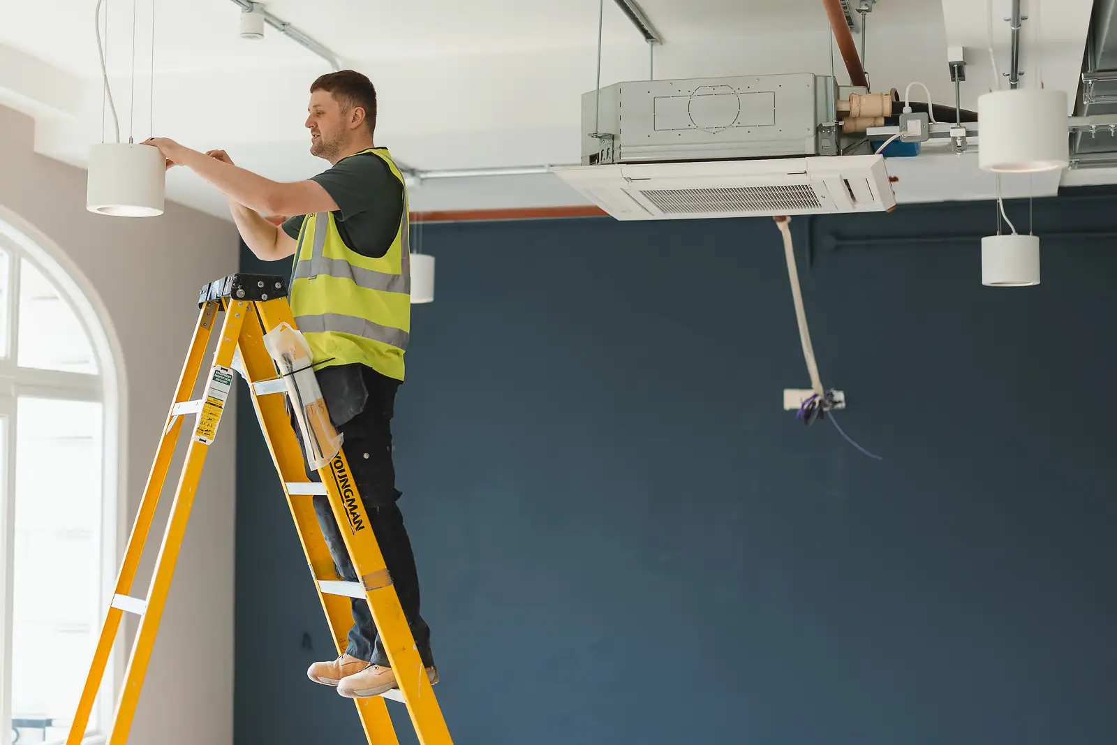 Electrician on a yellow ladder in profile, adjusting a pendant light fitting against a deep navy wall with arched window to the left