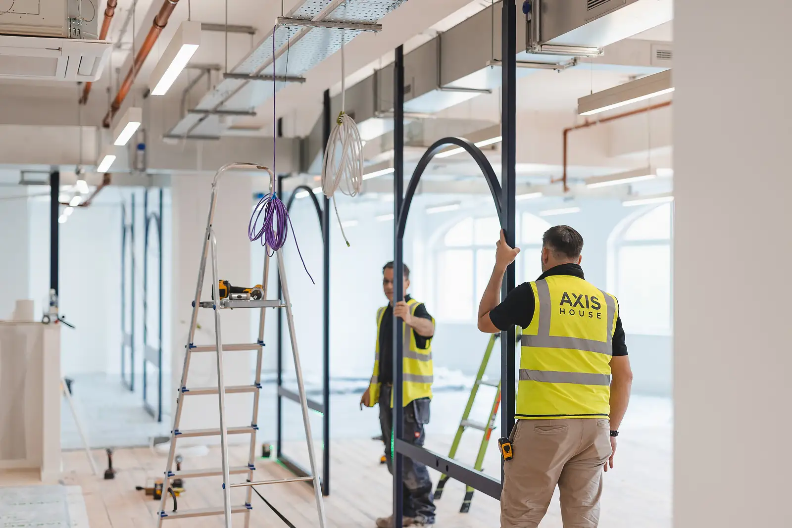 Two workers in branded hi-vis installing an arch frame along a long corridor, natural light flooding through at the far end