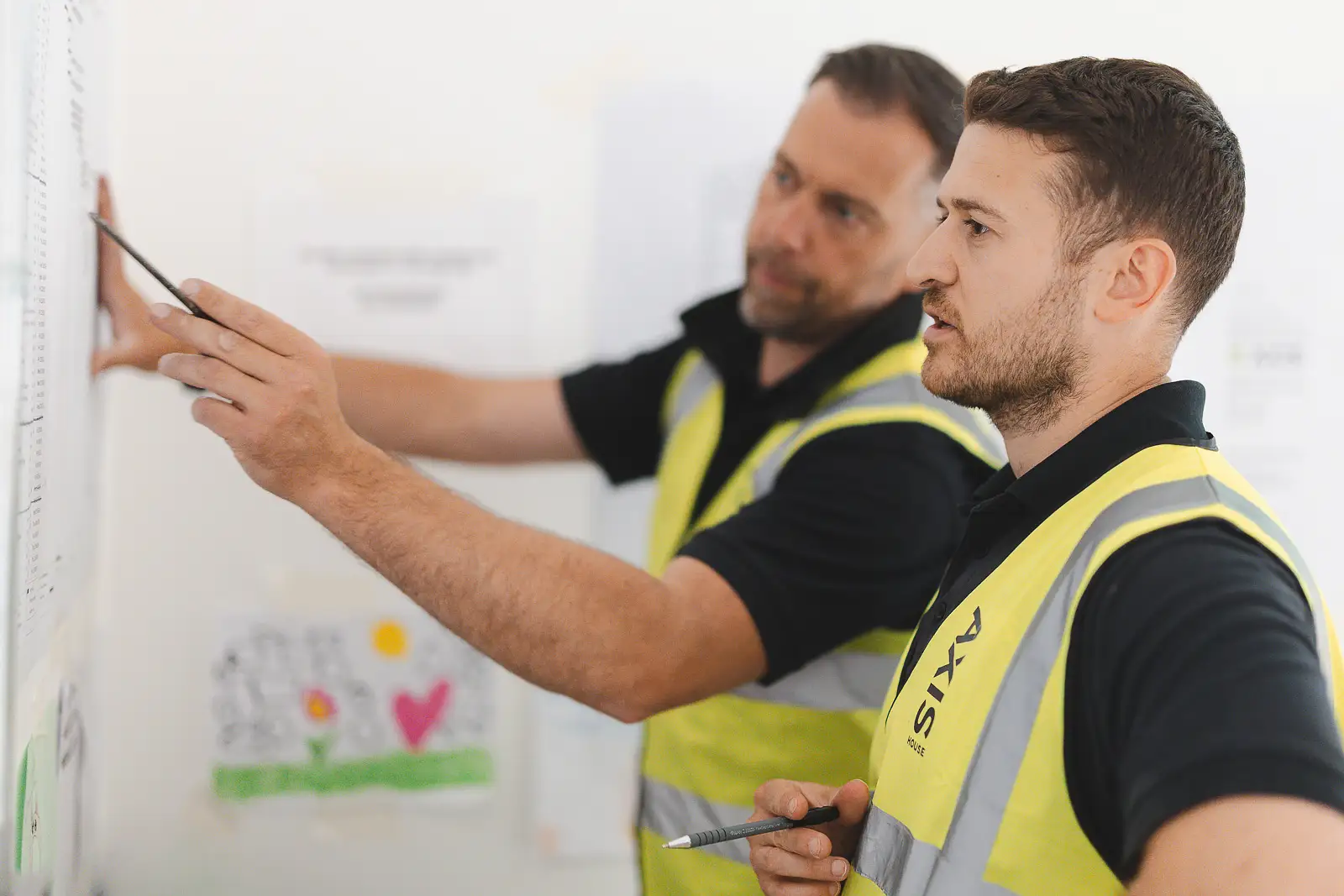 Tighter crop of two managers annotating site drawings pinned to the wall, younger manager in foreground with pen raised
