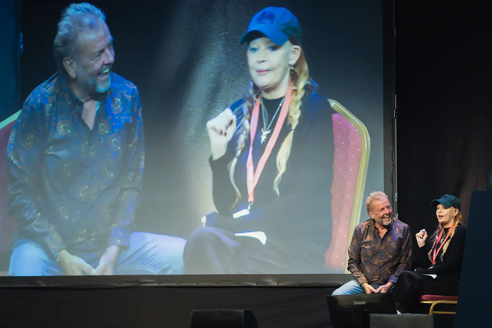 Two speakers in conversation on stage with their images projected on the large screen behind at a corporate event
