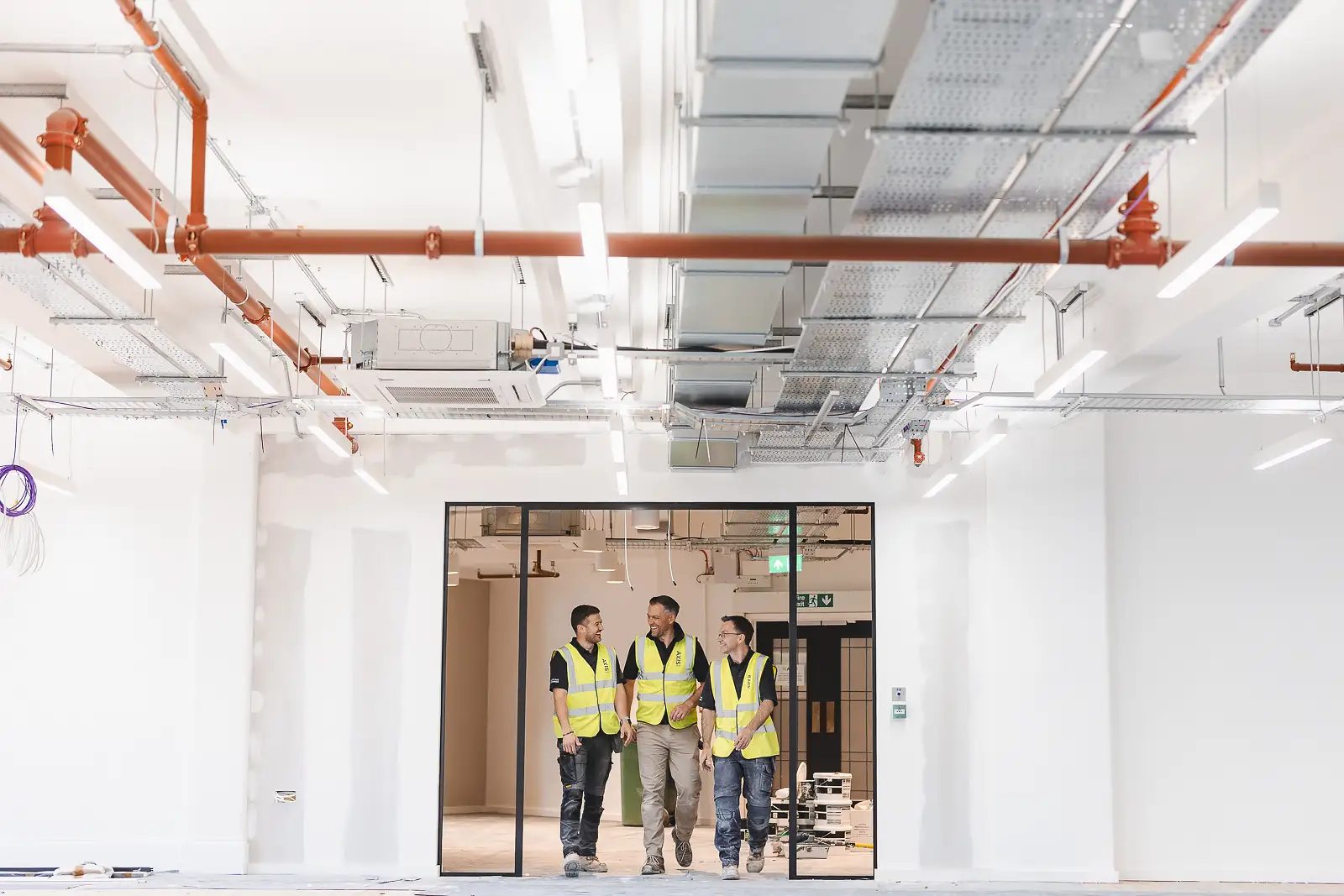 Wide-angle shot of three workers on a team walkthrough, exposed copper pipe services running across the ceiling above them