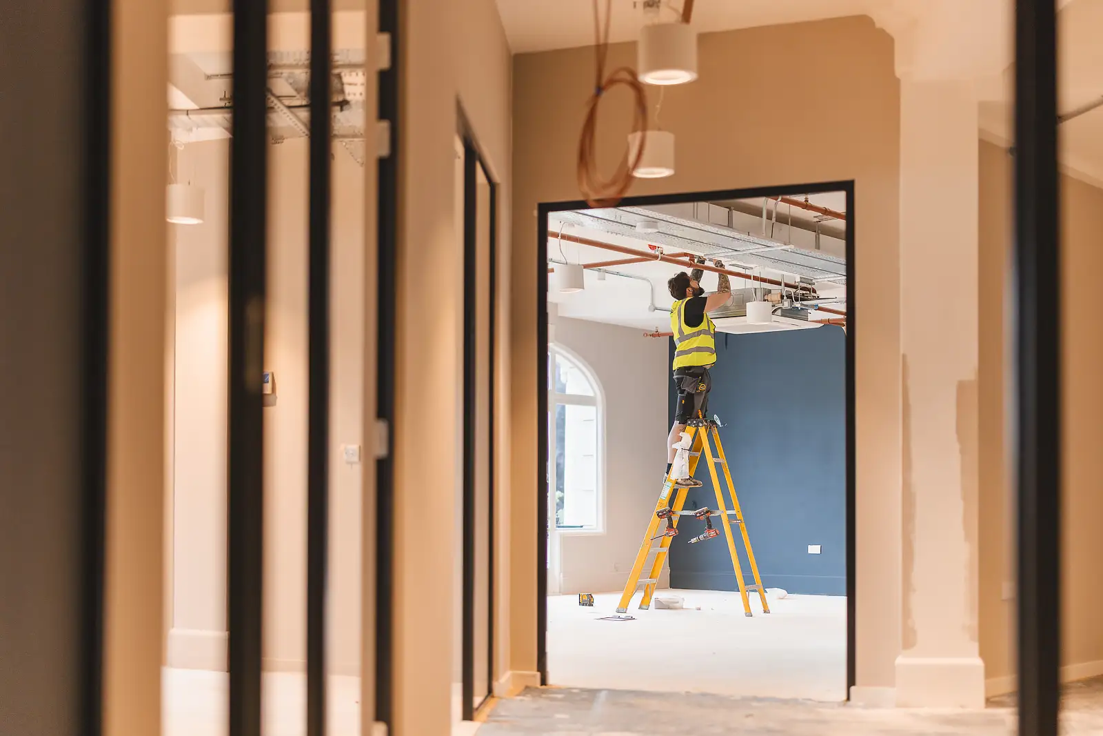 Worker positioned on a ladder at the far end of a long glass partition corridor, frames and doorways receding into the distance