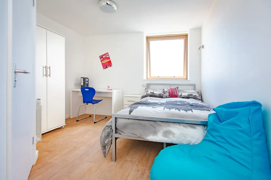 Bright accommodation bedroom with a desk, fitted storage and a turquoise beanbag beside the bed.