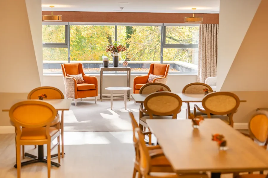 Communal dining area with round-backed chairs, tables and orange armchairs beside a wide window.