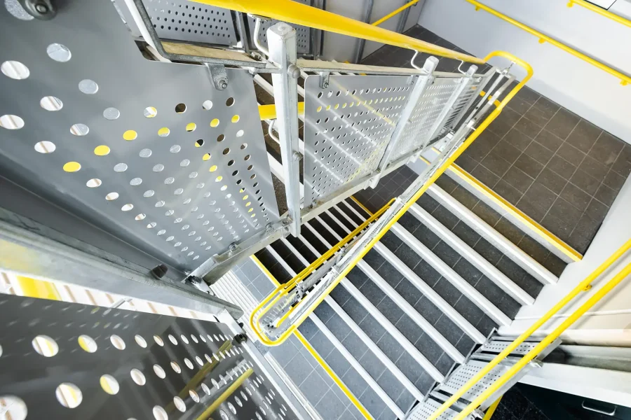 View down a metal stairwell, showing perforated panels, steel steps and yellow handrails.