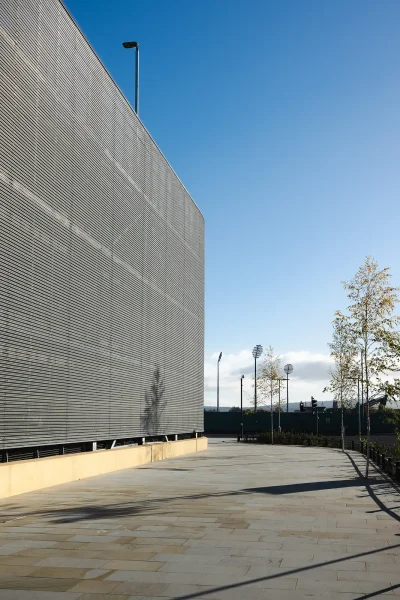 Vertical exterior view of metal facade panels beside a paved pedestrian route and young trees.