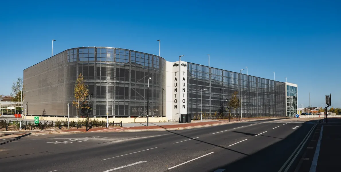 Wide exterior view of a multi-storey car park, showing its metal facade, glazed stair tower and roadside setting.