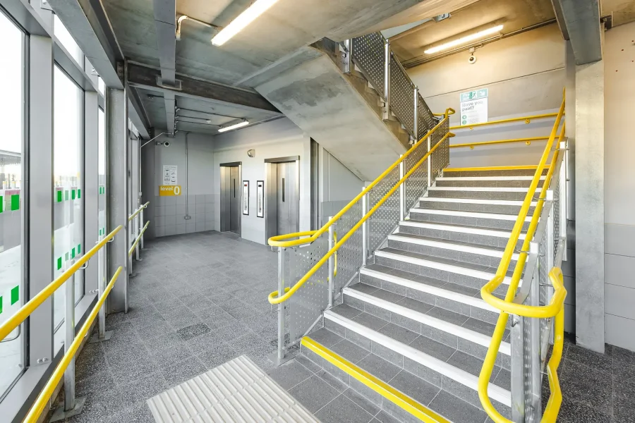 Car park entrance interior with a concrete staircase, yellow handrails, lifts and full-height glazing.