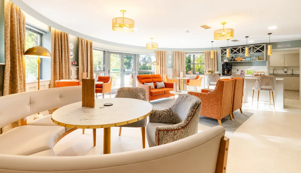 Bright communal lounge with curved booth seating, orange armchairs, round tables and a refreshment counter.