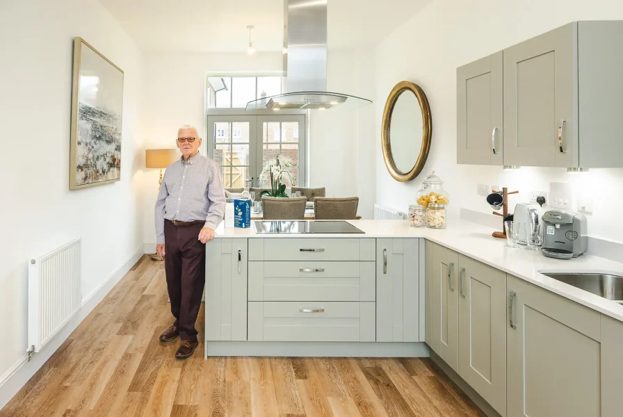 Man standing beside an island in a bright kitchen with grey cabinetry and wooden flooring.