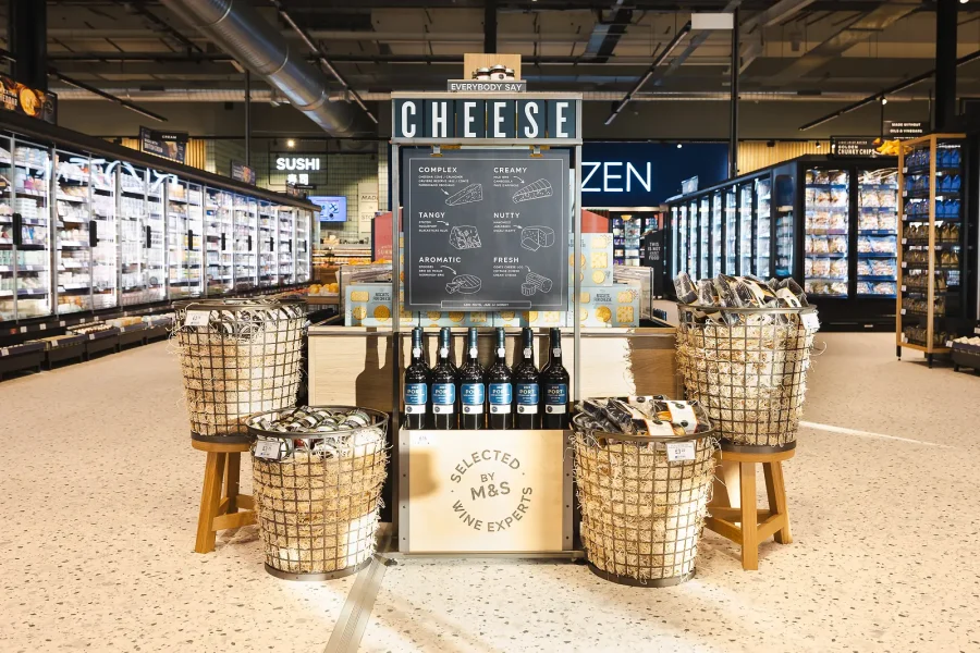 Cheese and wine arranged on a central display with wicker baskets, photographed as part of the retail brief.